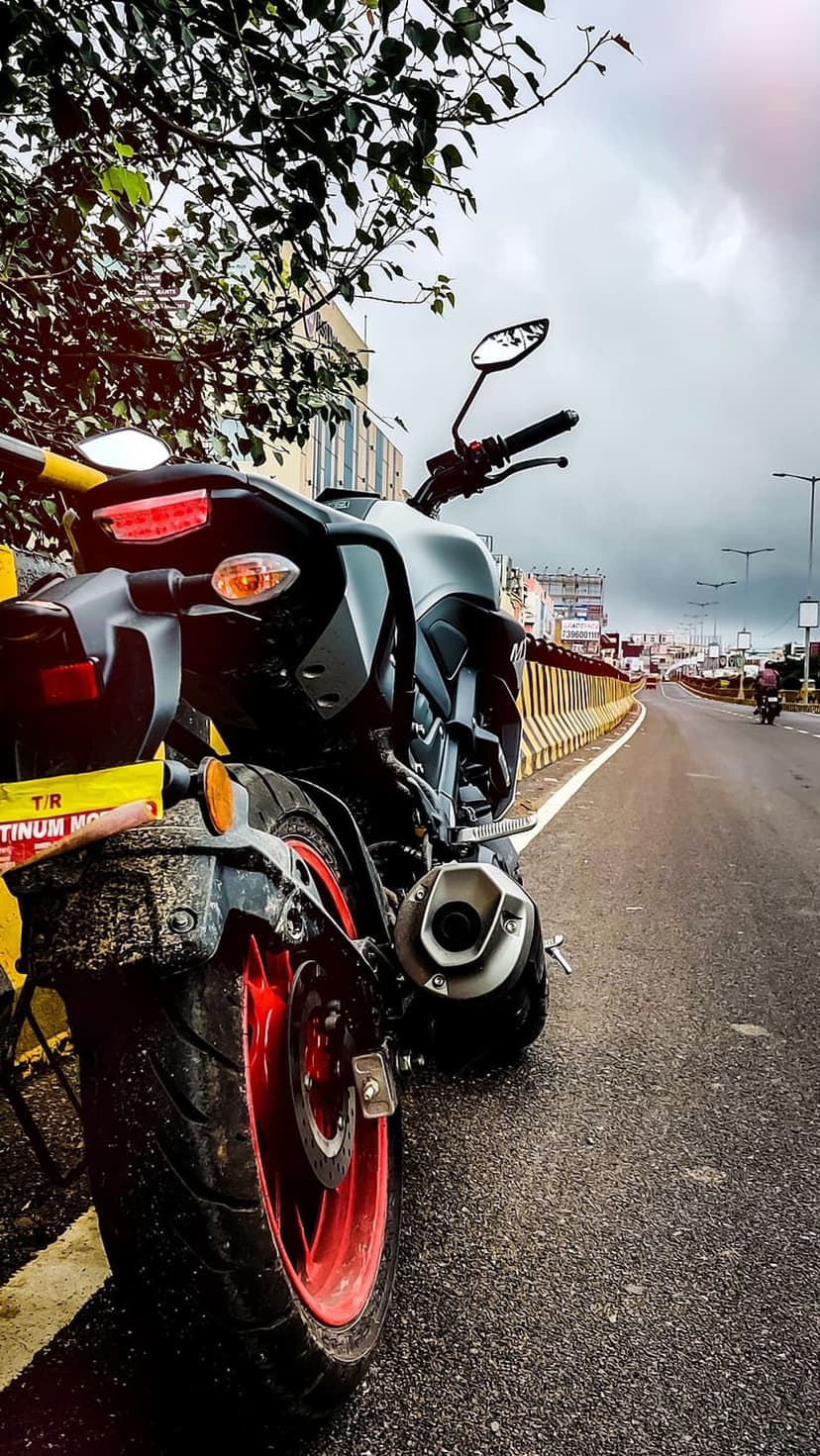 Black motorcycle rear tire with red rims on wet asphalt under an overcast sky with yellow road barriers.