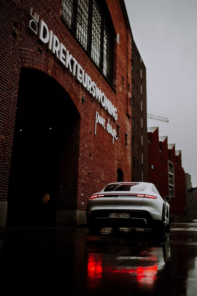 Silver Porsche Taycan with red taillights parked on a wet street in front of a historic red brick building.