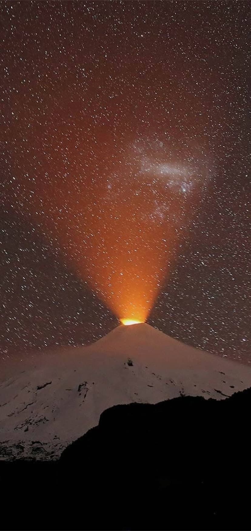 Snowy volcano peak emitting an orange light beam toward a starry night sky and the Milky Way nebula.