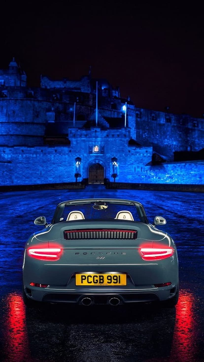 Gray Porsche 911 convertible parked on wet asphalt with red taillights glowing before a blue lit castle.
