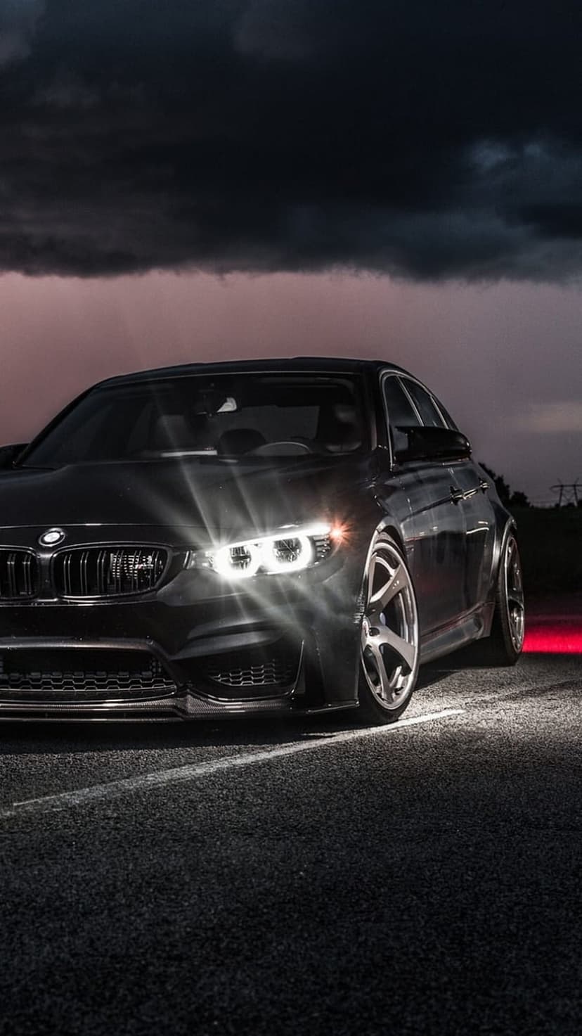 Black BMW M4 sports coupe with glowing LED headlights parked on a damp road under a stormy twilight sky.