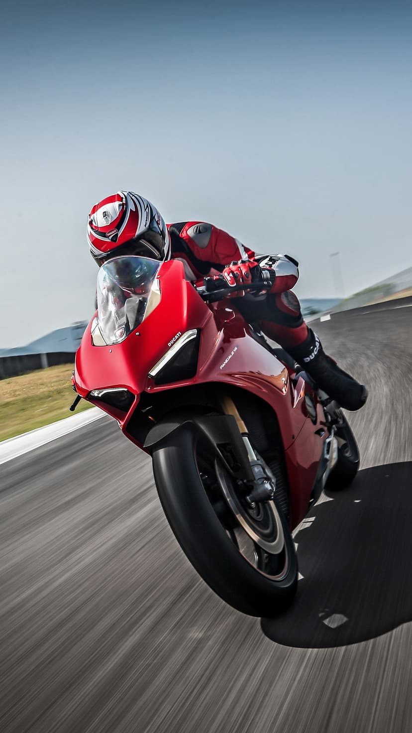 Ducati rider in red leathers leans low into a racetrack corner with motion blur under a clear blue sky.