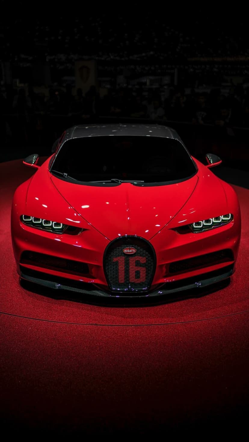 Red Bugatti Chiron with a number sixteen grille rests on a scarlet carpet under focused dramatic spotlighting.
