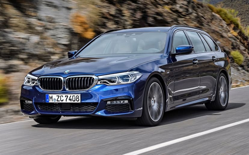 Dark blue BMW 5 Series Touring M Sport wagon driving fast on a mountain road with blurred rocky background.