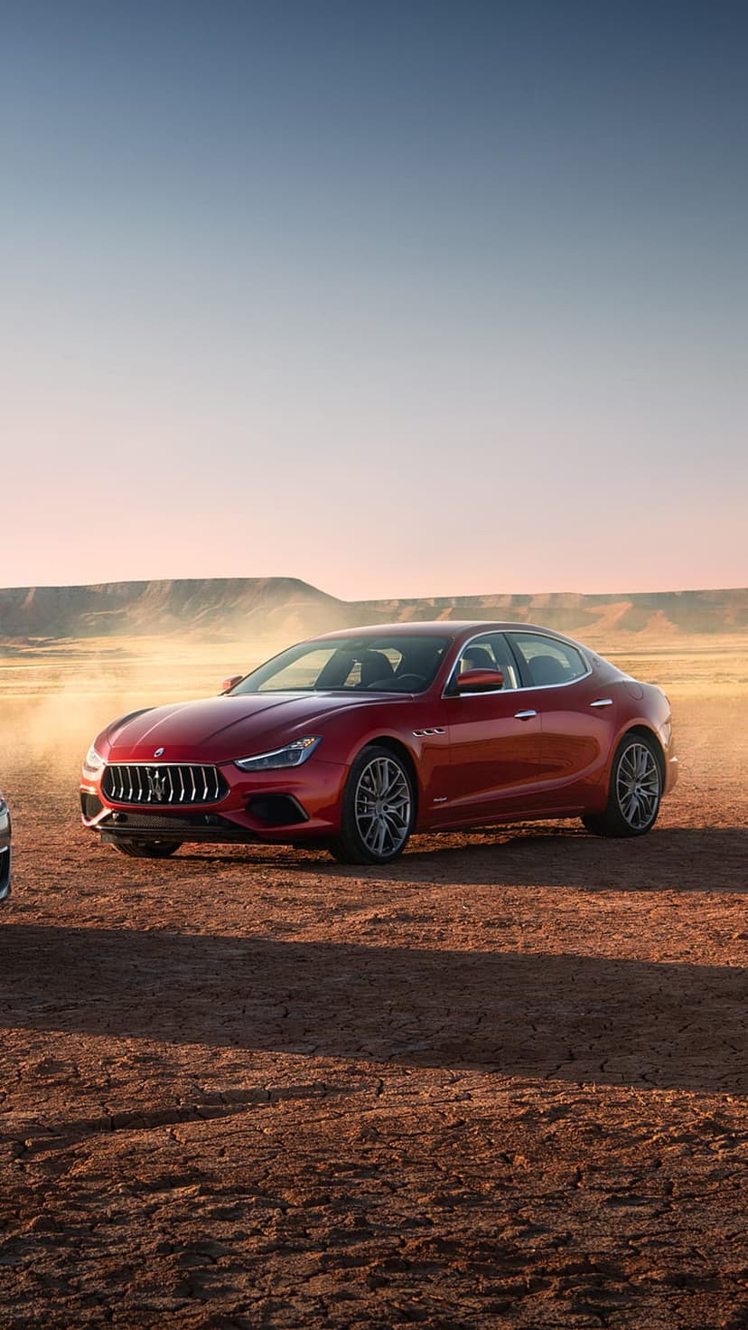 Red Maserati Ghibli sedan driving fast through a sandy desert with dust clouds swirling around the wheels.