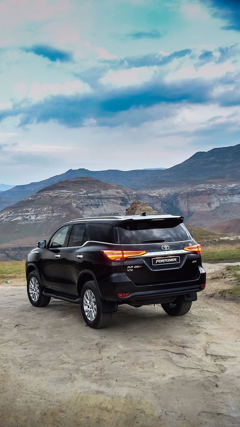 Black Toyota Fortuner SUV parked on a dirt path facing distant jagged mountains under a cloudy, overcast sky.