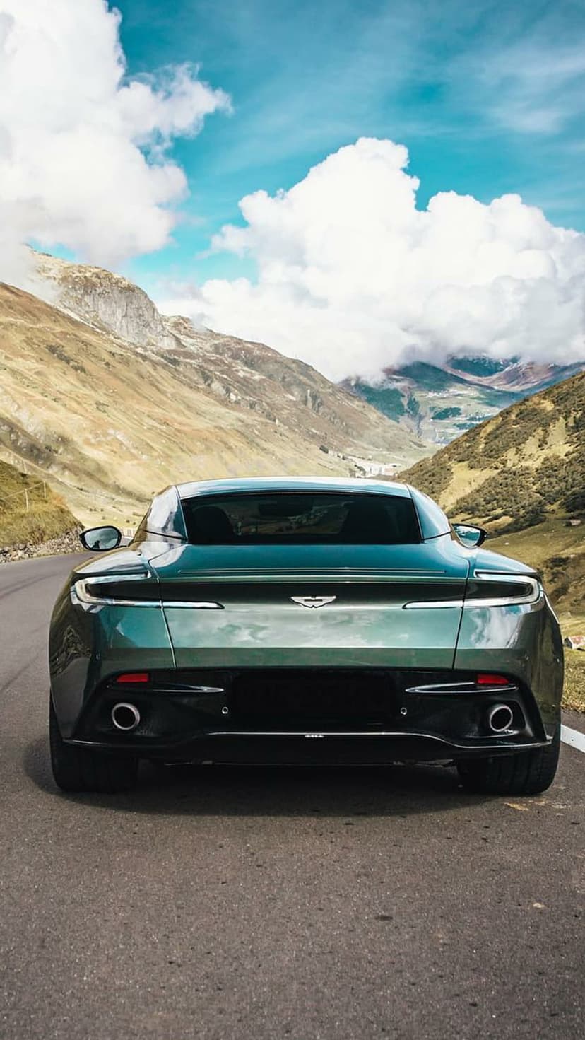 Aston Martin sports car rear view on a winding mountain road under a bright blue sky with fluffy white clouds.
