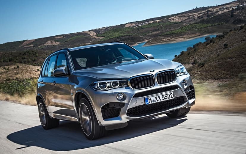 Silver BMW X5 M SUV driving on a winding road beside a blue lake and golden hills under a clear blue sky.