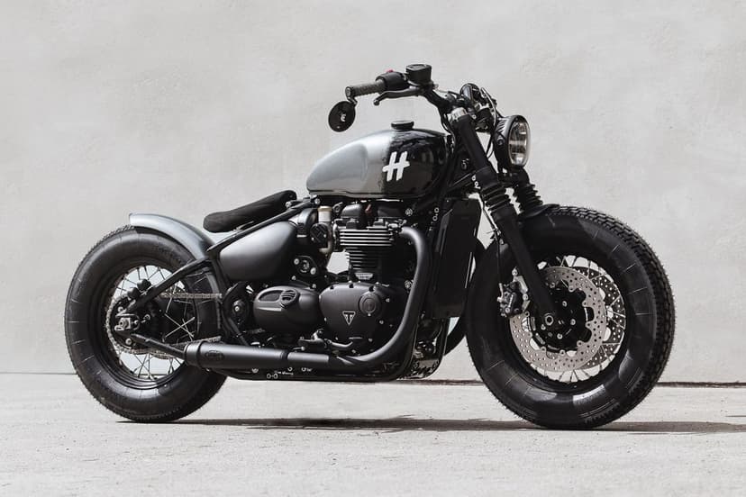 Custom black Triumph bobber motorcycle with grey tank parked against a minimalist white wall in bright light.