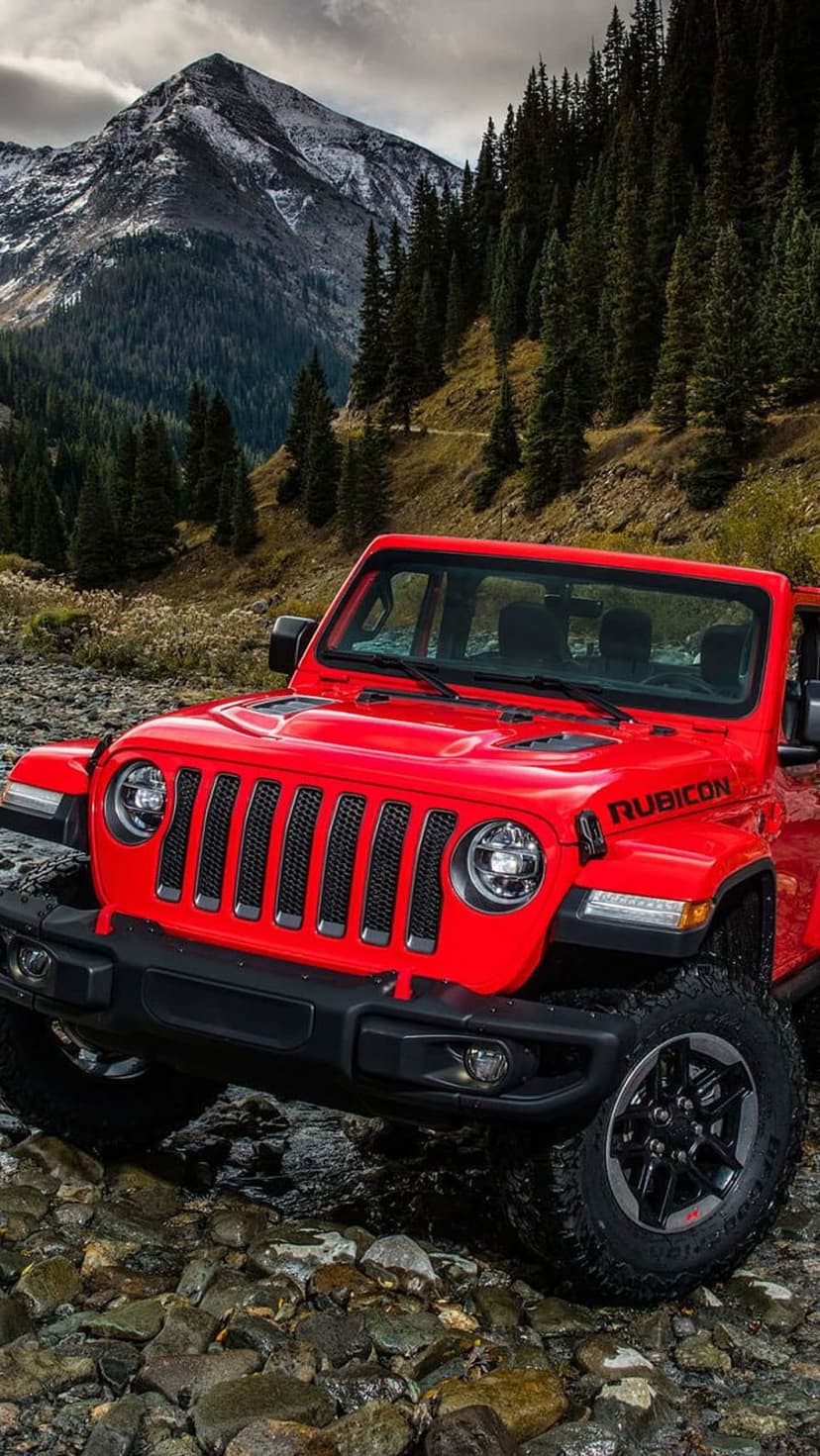Red Jeep Rubicon sits on a rocky riverbed with snow-capped mountains and a pine forest in the background.