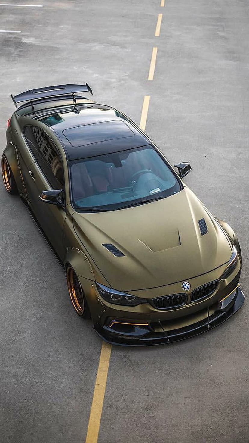 Gold BMW M4 coupe with widebody fenders, rear wing, and bronze wheels from an aerial drone perspective view.