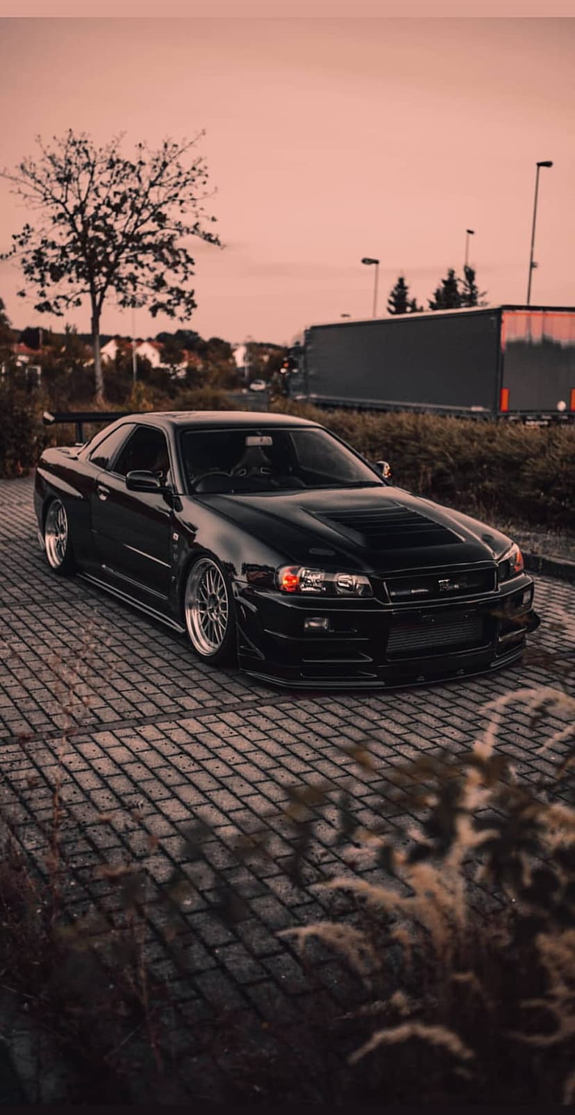 Black Nissan Skyline R34 GT-R with custom rims parked on paving stones under soft evening twilight lighting.