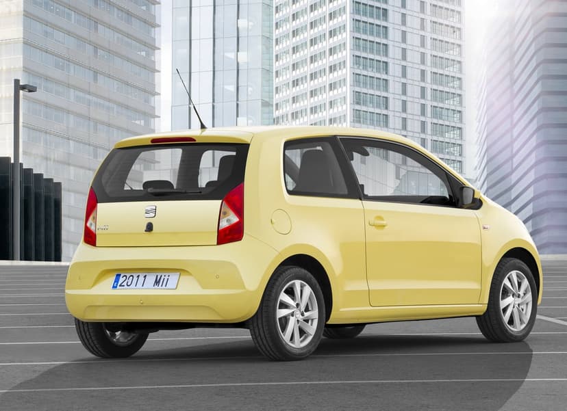 Rear view of a pale yellow SEAT Mii parked on an asphalt lot with glass skyscrapers in the background.