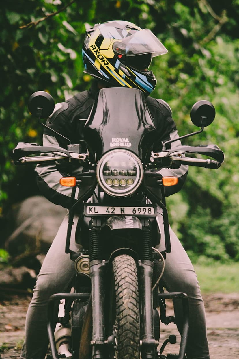 Motorcyclist in grey gear on a black Royal Enfield bike parked amidst vibrant green jungle plants and foliage.