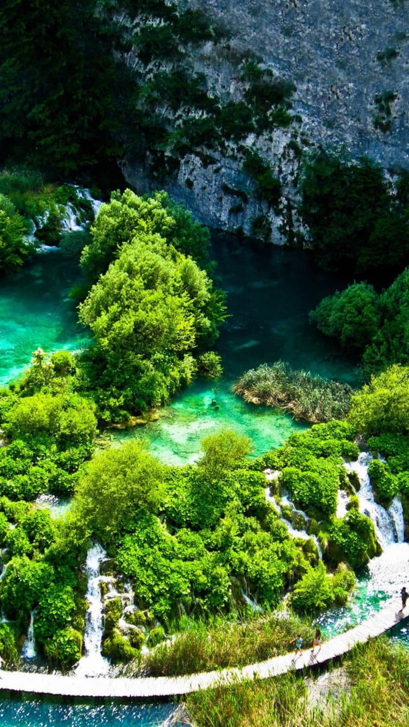 Turquoise Plitvice Lake waterfalls and a wooden boardwalk are viewed from above amidst lush green forest islands.