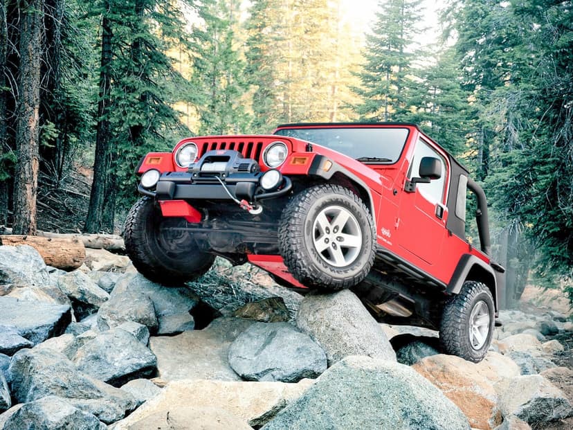 Red Jeep Wrangler TJ climbing over large gray rocks in a sunlit pine forest with its front tire elevated.