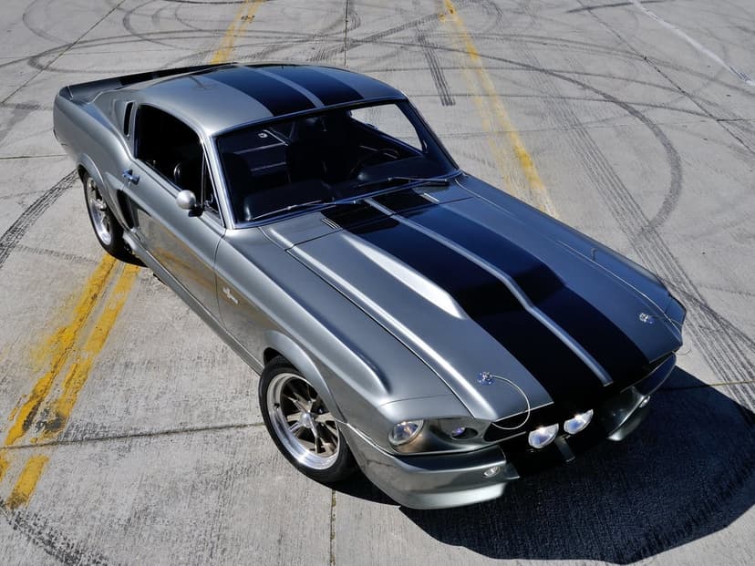 Silver 1967 Mustang Eleanor with black stripes on concrete pavement with tire marks and yellow lines.