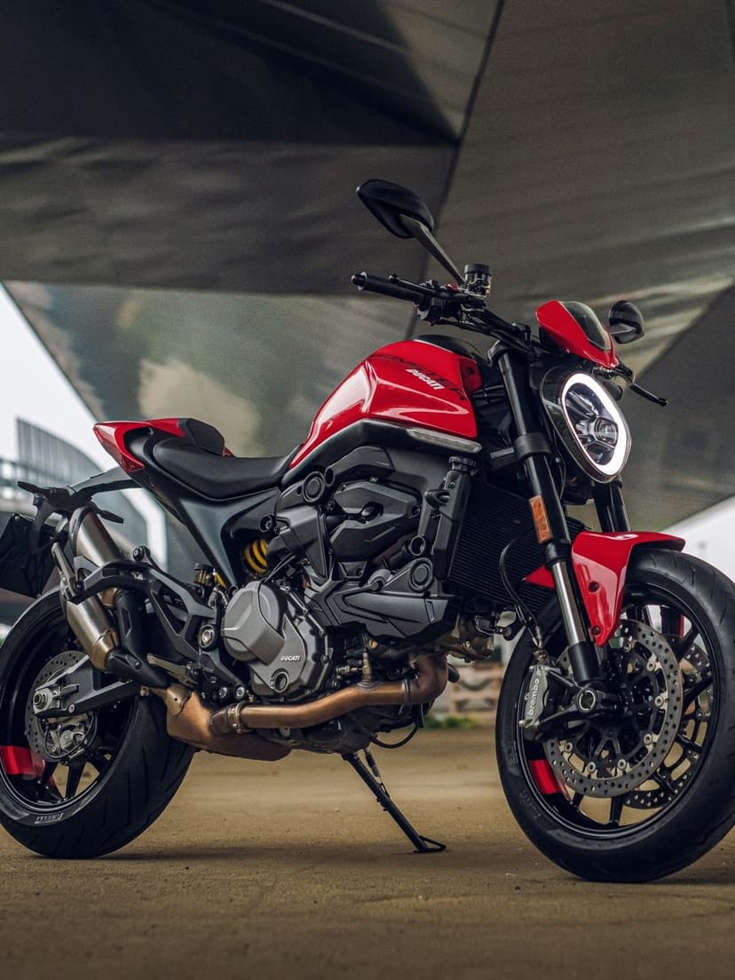 Red Ducati Monster motorcycle with a circular LED headlight parked under a concrete bridge in a portrait view.