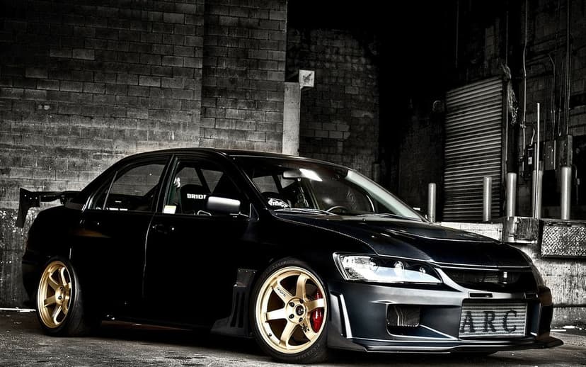 Black Mitsubishi Lancer Evolution with gold RAYS wheels and a carbon fiber hood in a dark brick warehouse.