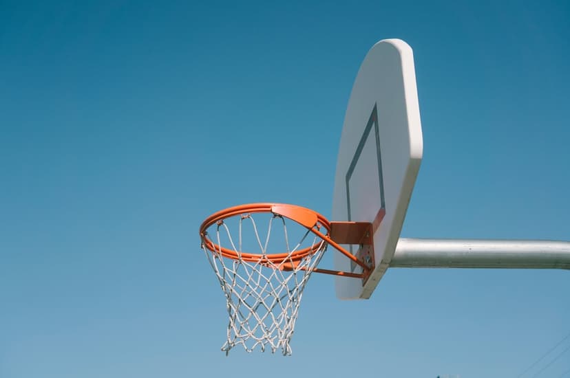 Basketball hoop backboard with an orange rim and white net positioned against a clear, solid blue summer sky.