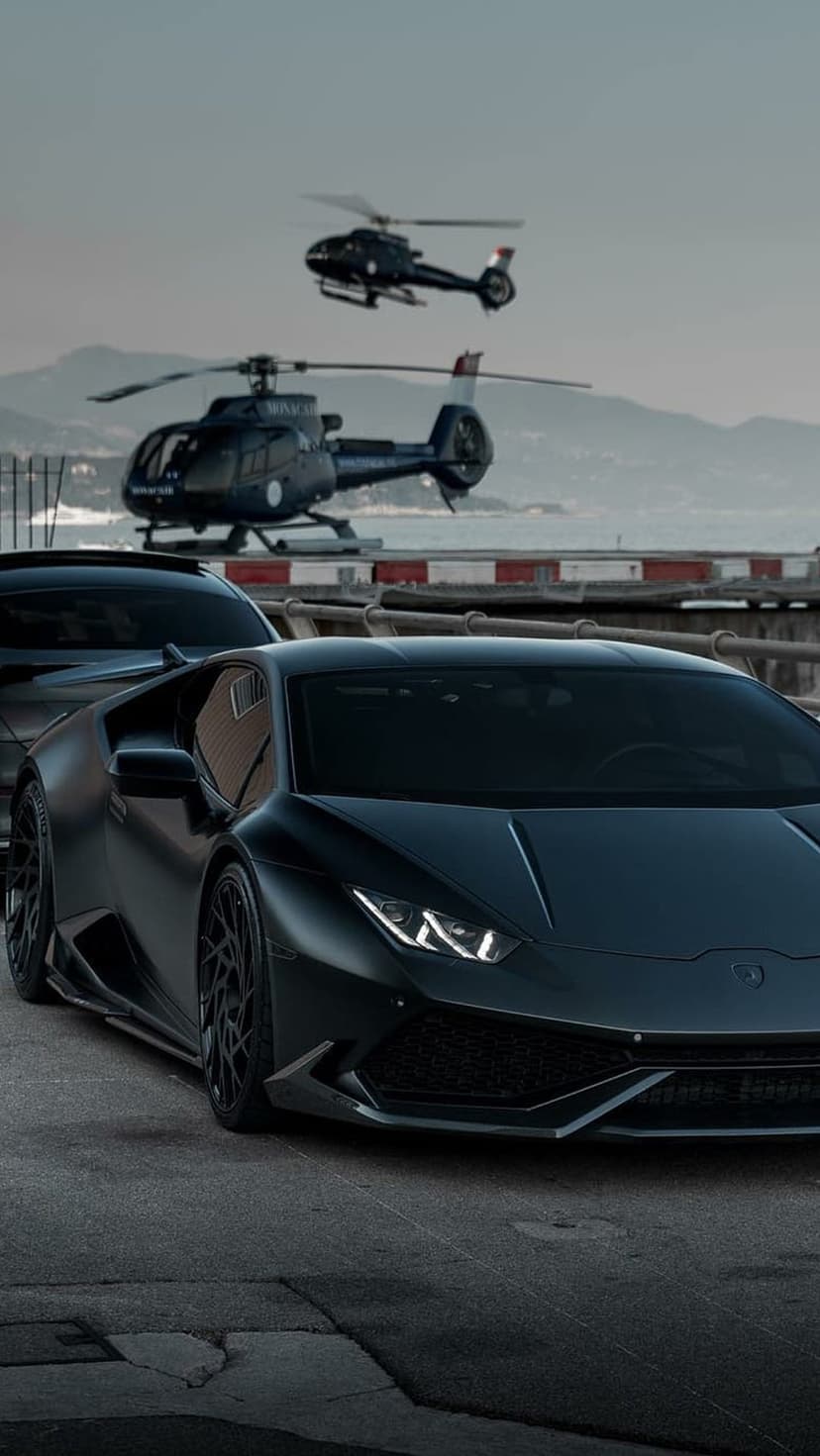Matte black Lamborghini supercar parked in front of two dark helicopters near a mountainous waterfront landscape.
