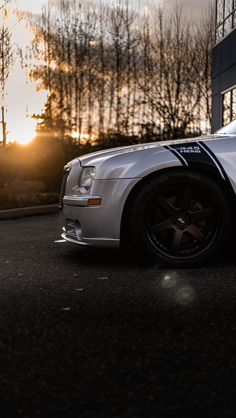 Silver Dodge Charger 345 Hemi with black racing stripes parked on asphalt during a warm golden hour sunset glow.