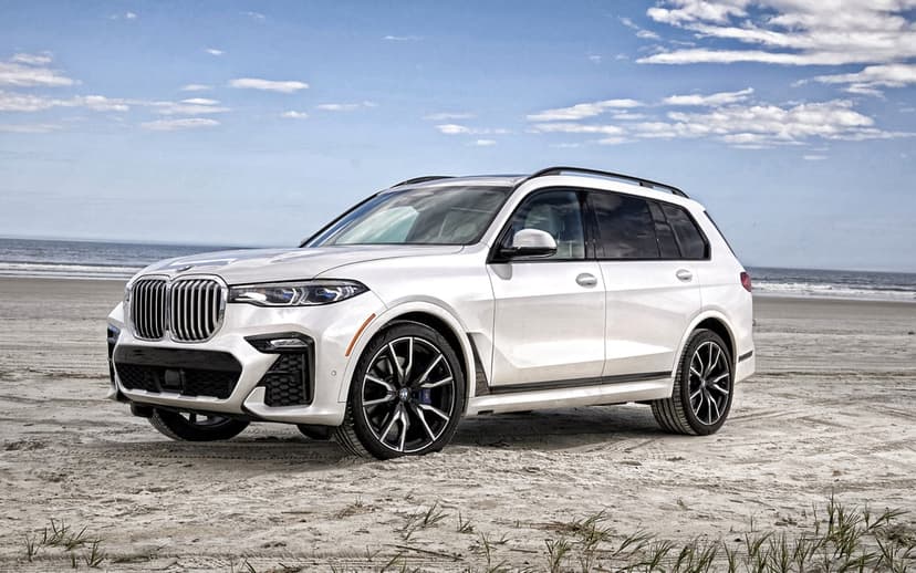 White BMW X7 SUV parked on a sandy beach with tire tracks, overlooking the blue ocean under a bright sky.