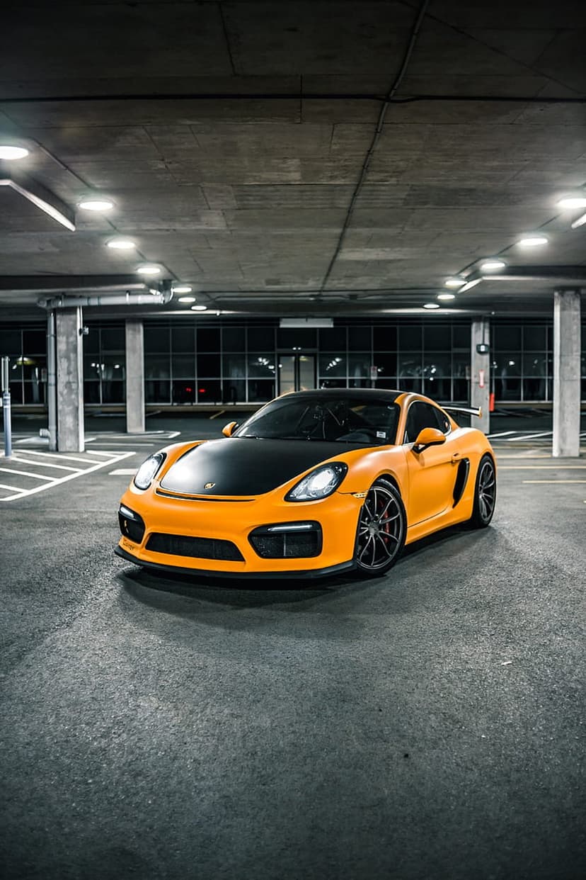 Orange Porsche Cayman GT4 with a black hood parked on a concrete floor in a dimly lit modern parking garage.