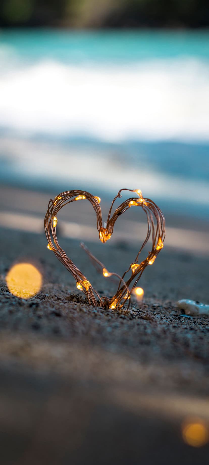 Heart shaped fairy lights glowing on wet sand at sunset with a blurred ocean background and golden horizon.