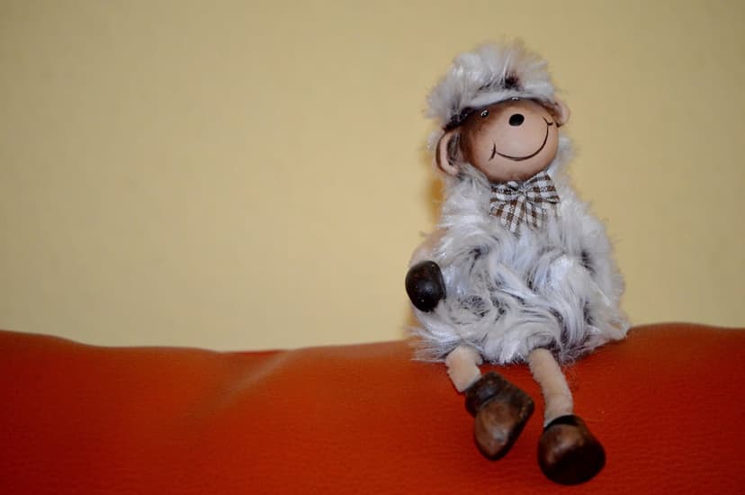 Fluffy grey sheep plush toy with a checkered bowtie sits on a vibrant orange sofa against a soft yellow wall.
