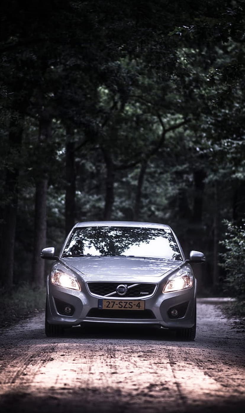 Silver Volvo C30 hatchback with lit headlights driving on a gravel road through a dense, dark pine forest.