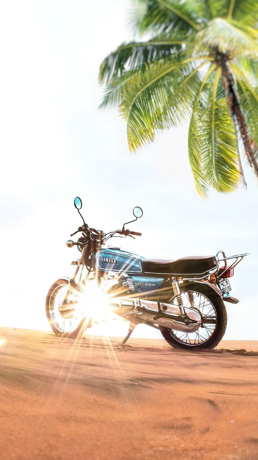 Blue Yamaha RX100 motorcycle parked on white sand under a green palm tree with a bright golden lens flare.