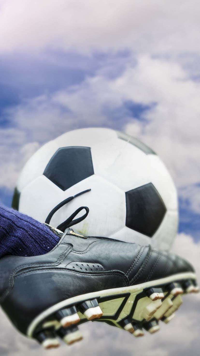 Black leather soccer cleat with visible studs resting on a black and white ball under a soft blue cloudy sky.