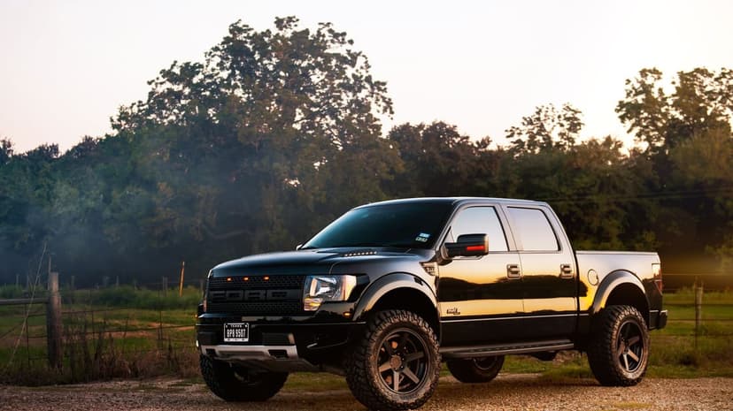 Black Ford F-150 Raptor pickup truck with off-road tires on a gravel road at sunset with golden reflections.