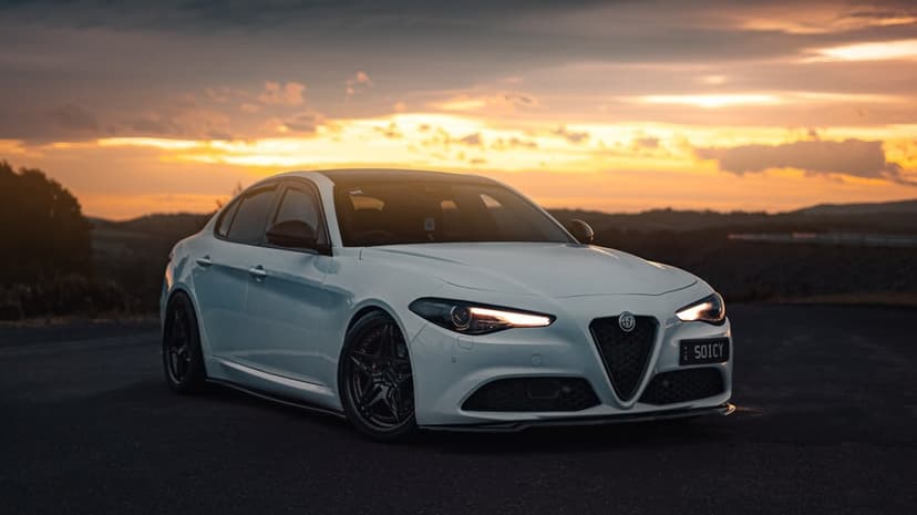 White Alfa Romeo Giulia with black wheels on a road during a golden hour sunset with a cloudy sky.