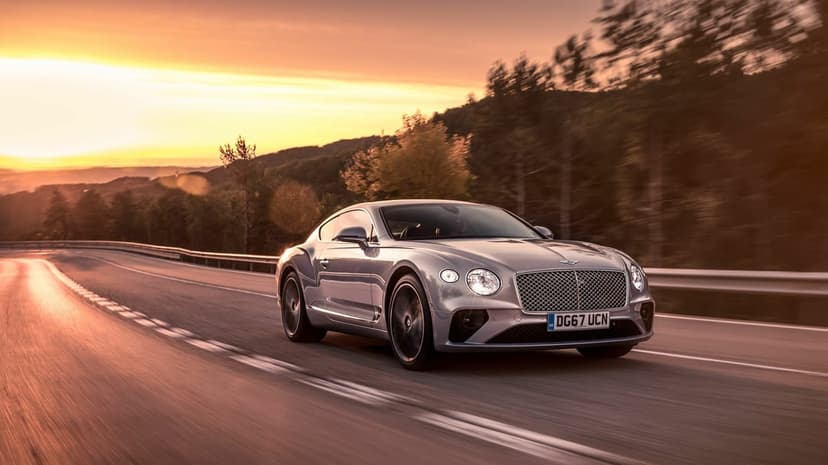 Silver Bentley Continental GT driving on a winding forest road during a golden sunset with motion blur.