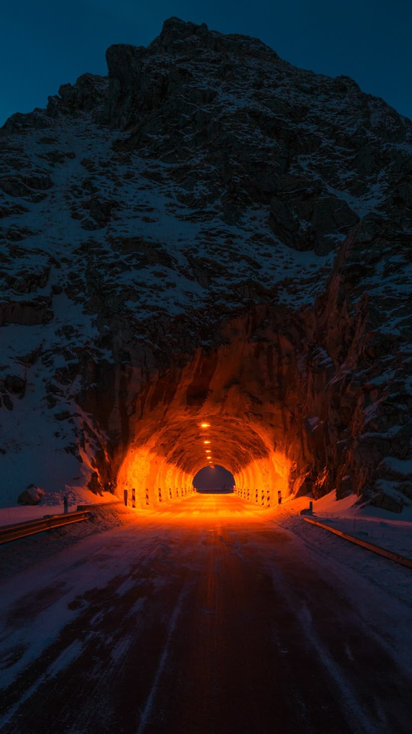 Orange-lit tunnel opening at the base of a jagged, snow-covered mountain peak under a dark blue twilight sky.