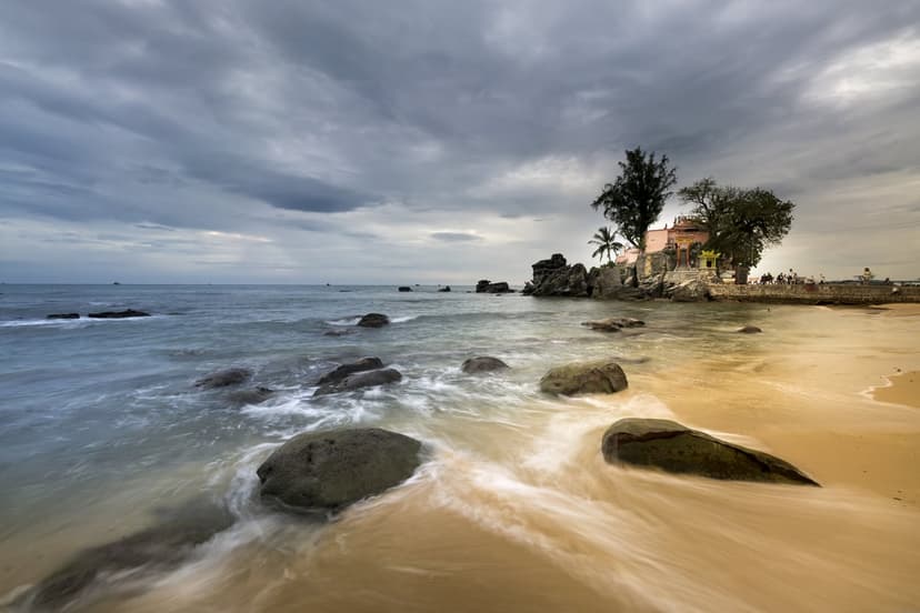 Small island shrine on jagged rocks above misty waves and a sandy shore under a dark, swirling stormy sky.