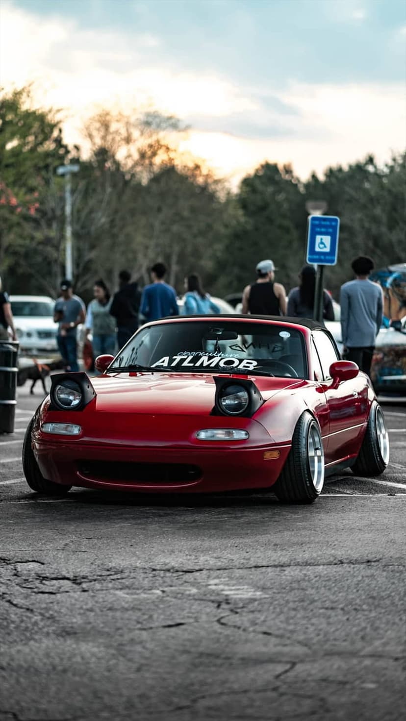 Red Mazda Miata with pop-up headlights raised, parked at an outdoor car meet during a golden hour sunset.