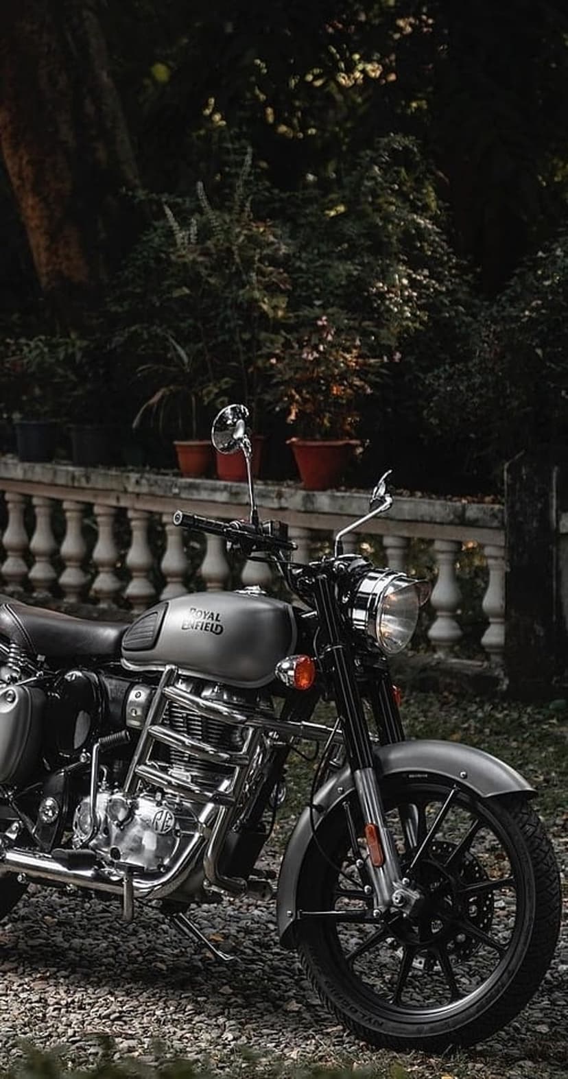 Grey Royal Enfield motorcycle on a gravel path beside stone railings and dense green garden bushes in soft light.