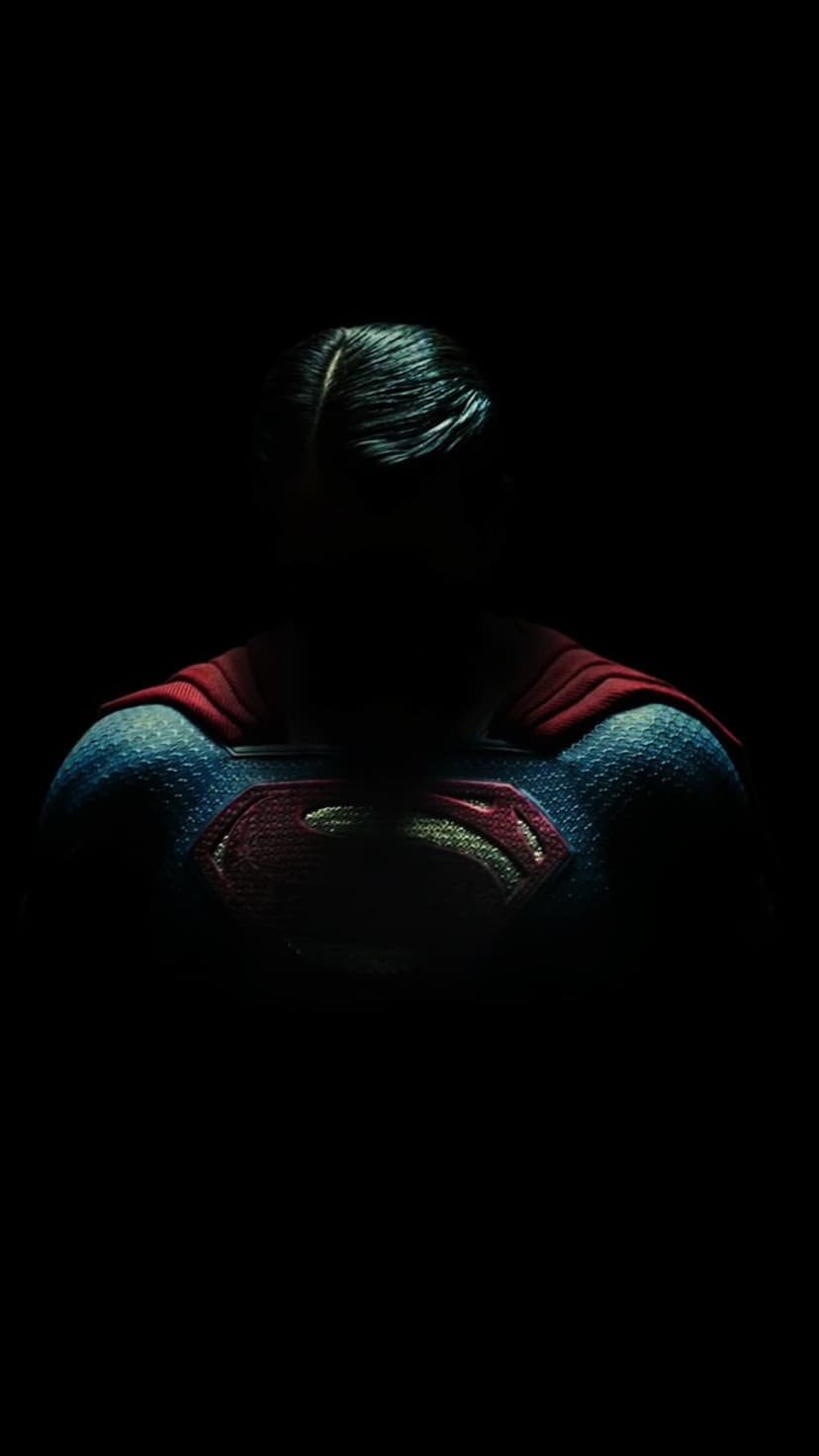 Superman's raised 'S' emblem and blue textured suit fabric emerge from a pitch-black background with red cape.