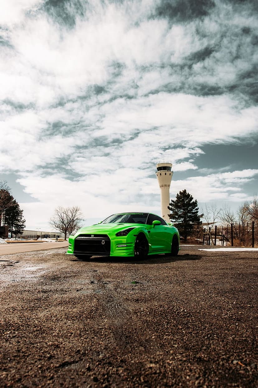 Neon green Nissan GT-R sports car parked on gravel with an airport control tower under a dark cloudy sky.