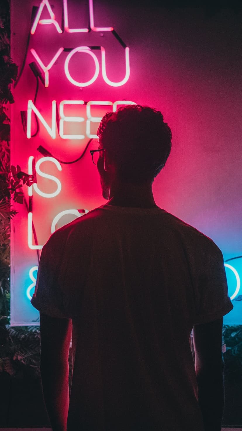 Silhouetted person standing before a glowing pink neon sign reading All You Need Is Love in a dark room.