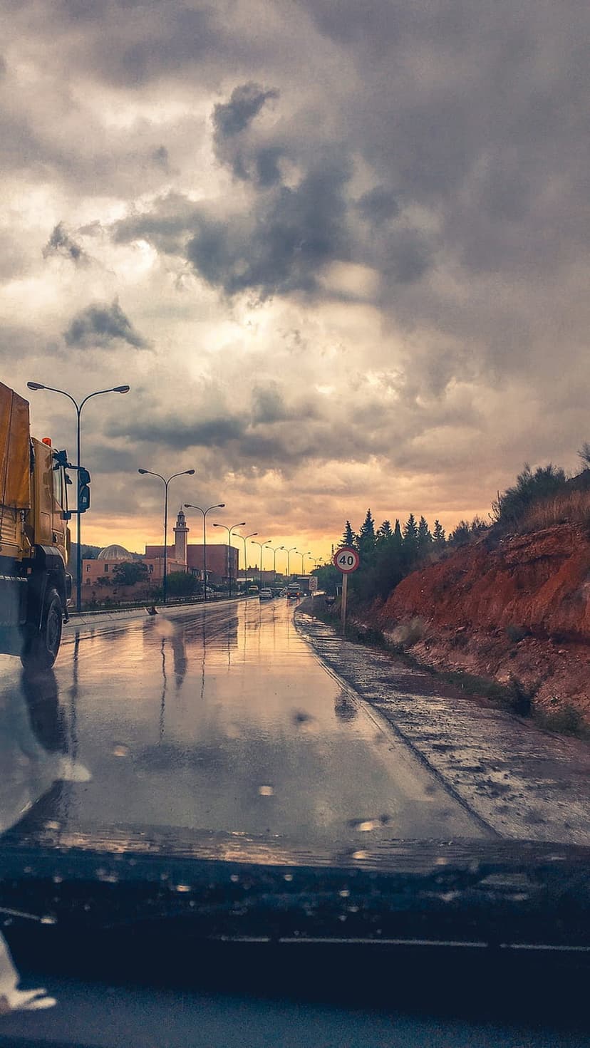 Wet highway road reflecting a golden sunset with a truck and a mosque minaret in the distance under dark clouds.