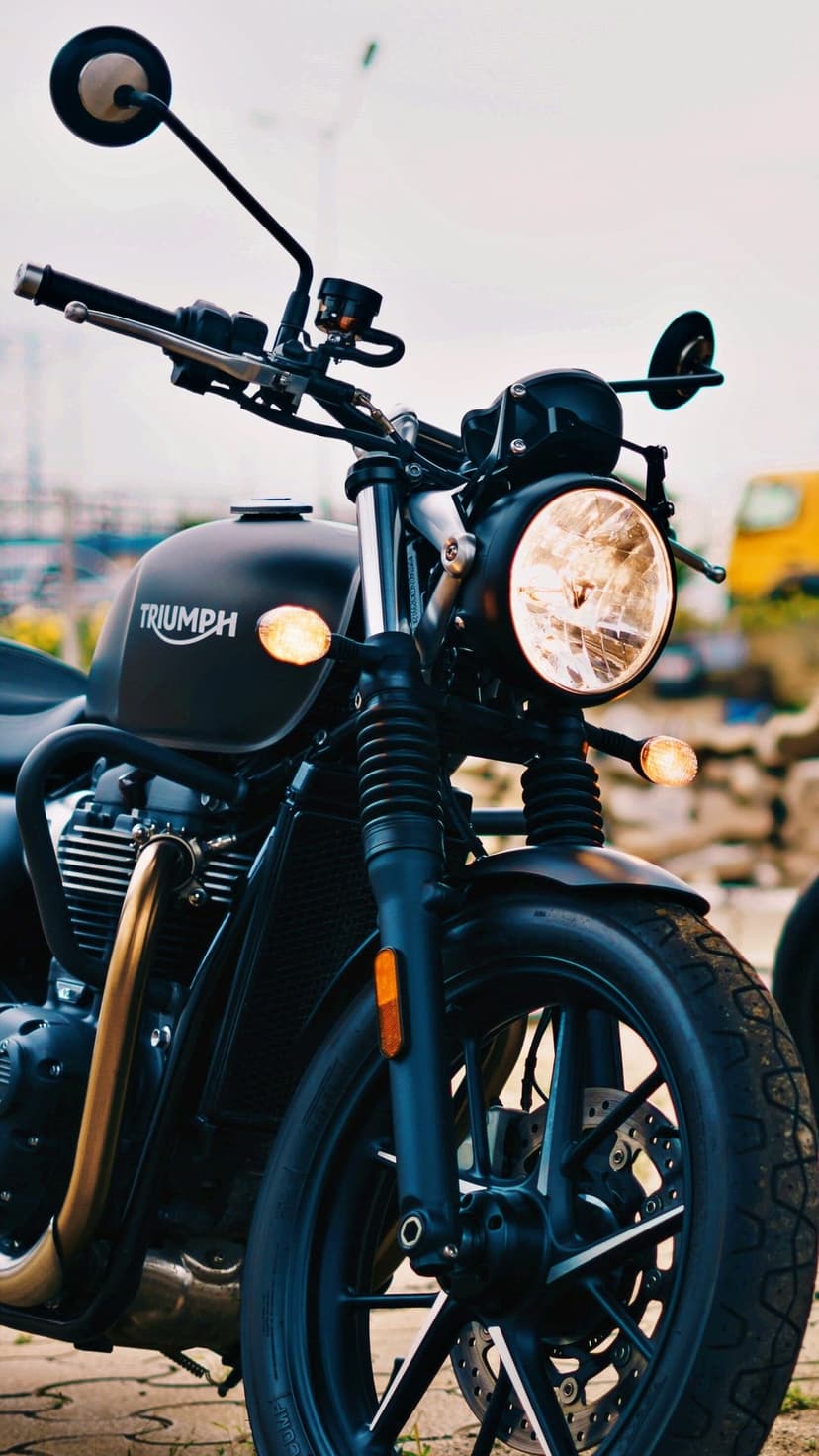 Close-up of a matte black Triumph motorcycle with a glowing circular headlight and gold exhaust pipe.