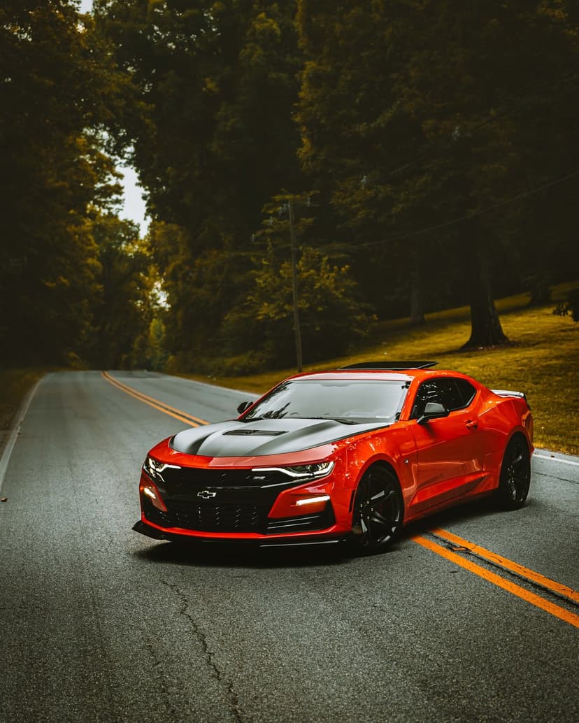 Red Chevrolet Camaro drives down an empty asphalt road bordered by green trees and golden roadside grass.
