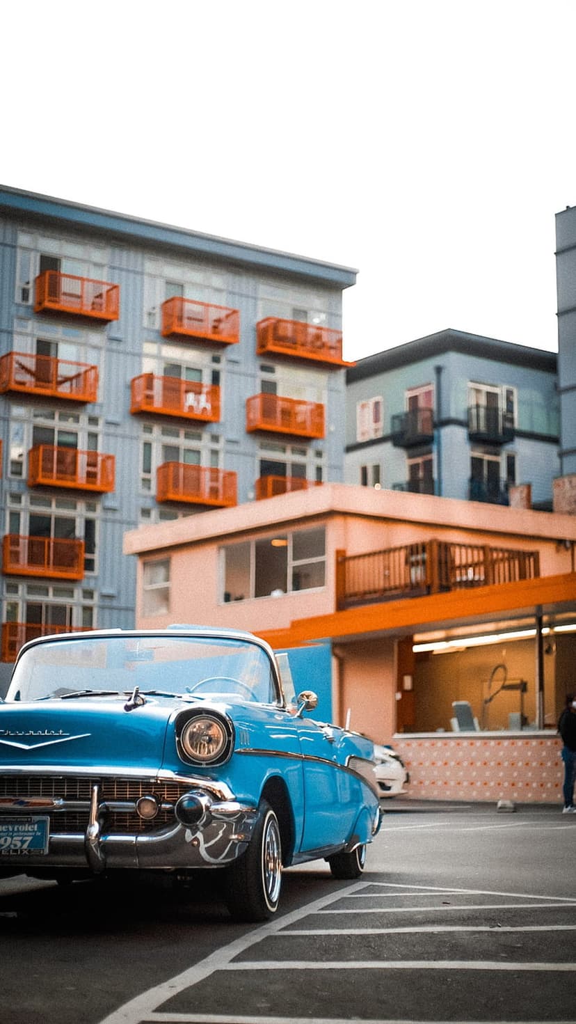 Blue Chevrolet Bel Air convertible parked in front of modern gray apartment buildings with orange balconies.