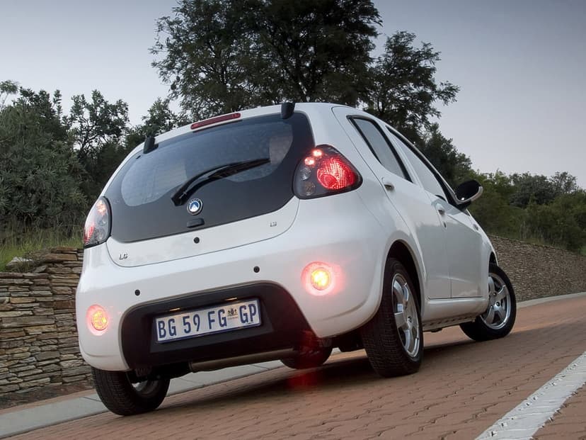 White Geely LC hatchback from a low rear angle on a brick road with lit taillights and a tree background.