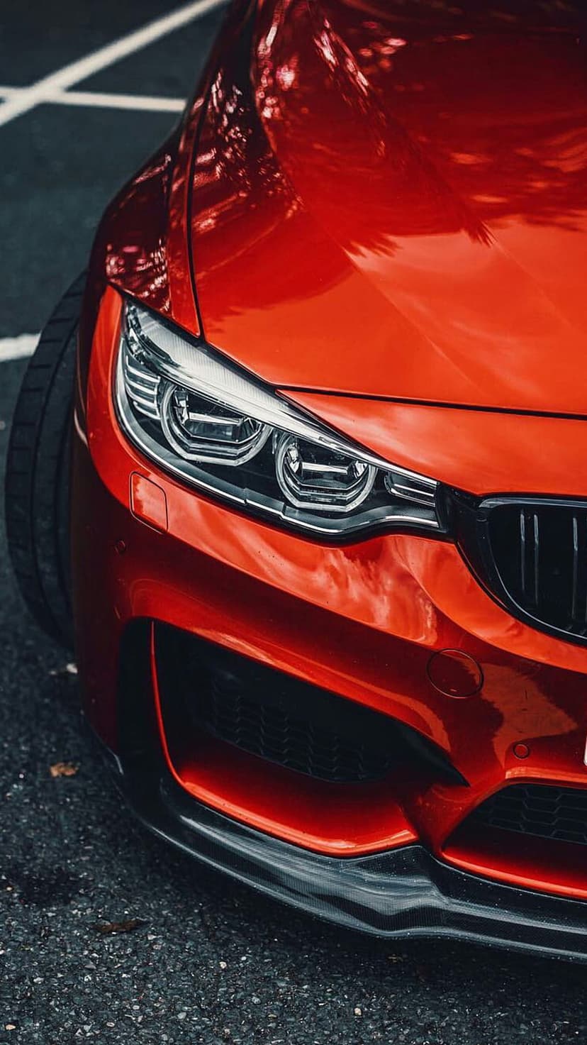 Red BMW M4 LED headlight and aggressive front bumper assembly shown from a low angle on textured dark asphalt.