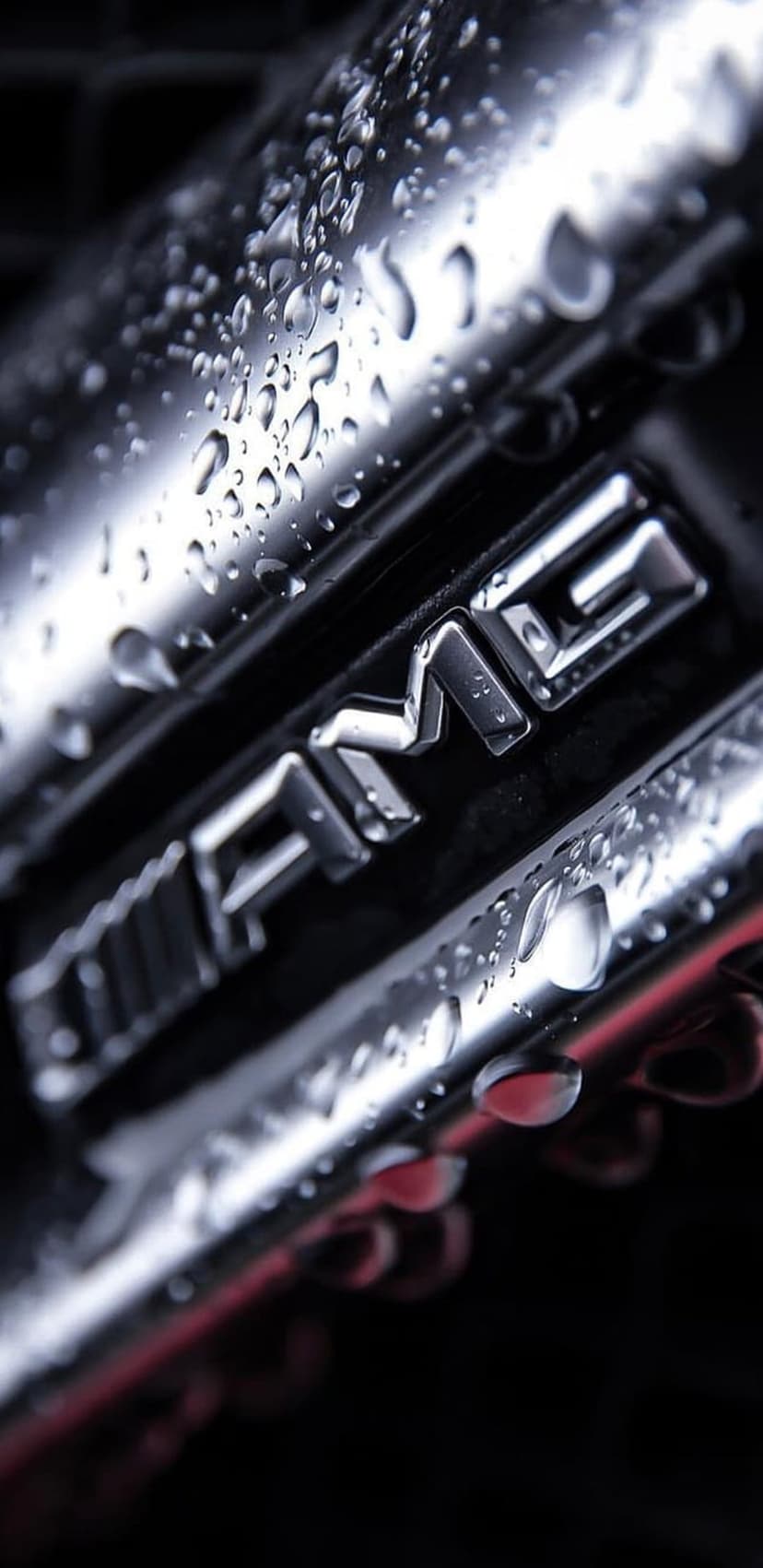 Chrome AMG badge covered in clear water droplets against a dark blurred background on a luxury car exterior.
